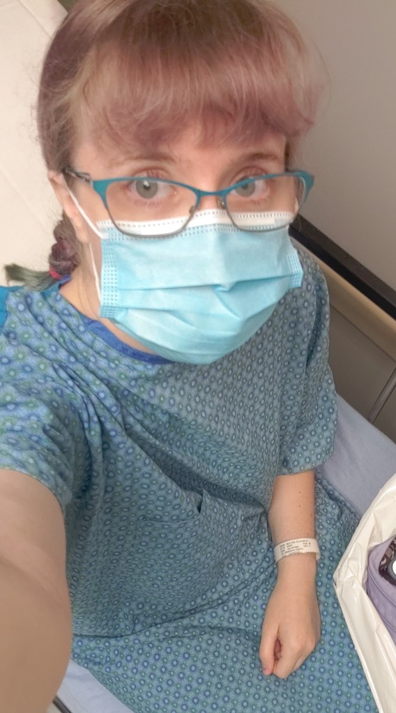 White woman with a blue surgical mask, blue glasses, and a blue hospital gown sitting on a hospital bed.